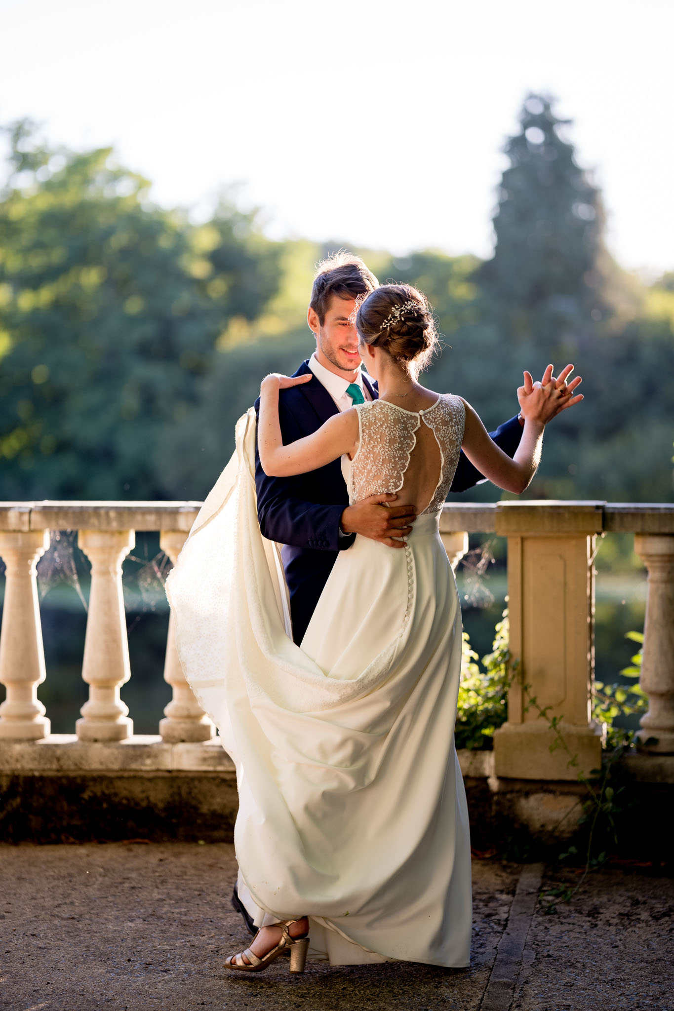 A couple dances together on an outdoor stone terrace with a classical balustrade railing, with a body of water and trees visible in the background. The bride wears an ivory sleeveless gown with a beaded lace illusion back featuring a scalloped neckline, button detailing down the spine, and a flowing chiffon skirt; her hair is styled in an updo adorned with a delicate gold leaf hairpiece, and she wears gold block-heel sandals. The groom wears a navy suit with a teal tie and white dress shirt, and he is smiling as he holds the bride in a dance position, lifting the skirt of her dress. The shot is a full-length portrait taken in warm golden-hour light, with the bride's back to the camera and the groom's face visible over her shoulder.