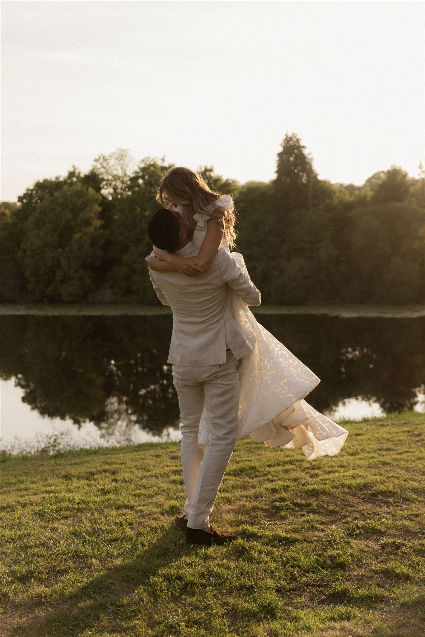 An outdoor couple portrait taken during golden hour beside a calm lake. The groom, wearing a light beige linen suit with brown loafers, is lifting the bride off the ground as she wraps her arms around his neck. The bride wears a white textured or lace gown with a flowing skirt that catches the warm light. Both face each other with their backs largely to the camera. The setting is a grassy lakeside with tree-lined banks reflected in the still water. The warm, low-angle sunlight casts a golden tone across the entire scene. The composition is a full-length portrait shot from behind.