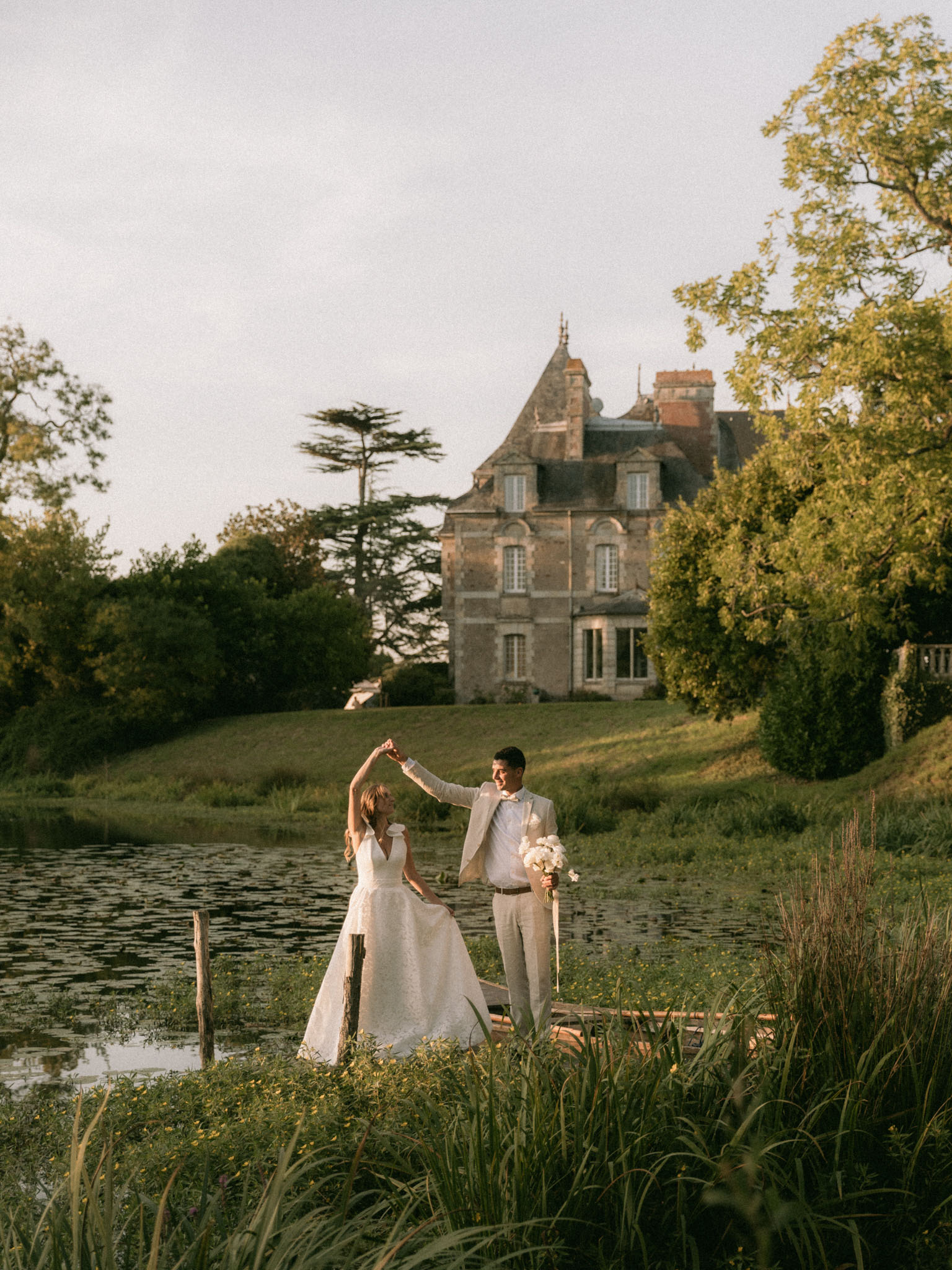 A couple portrait taken outdoors at golden hour beside a pond on the grounds of a French château. The groom, wearing a light beige suit with a bow tie, spins the bride by raising her hand while holding her white bouquet — composed of what appear to be white peonies or ranunculus — in his other hand. The bride wears a white A-line gown with a deep V-neckline and lace or textured fabric detail, and the two are standing on or near a small wooden dock at the water's edge. The château, a multi-story stone manor with steep slate roofing and pointed turrets, is visible in the background across the pond. The overall styling leans classic with a relaxed, romantic feel. Wide portrait shot with the venue prominently featured in the background. Potential venue feature image.