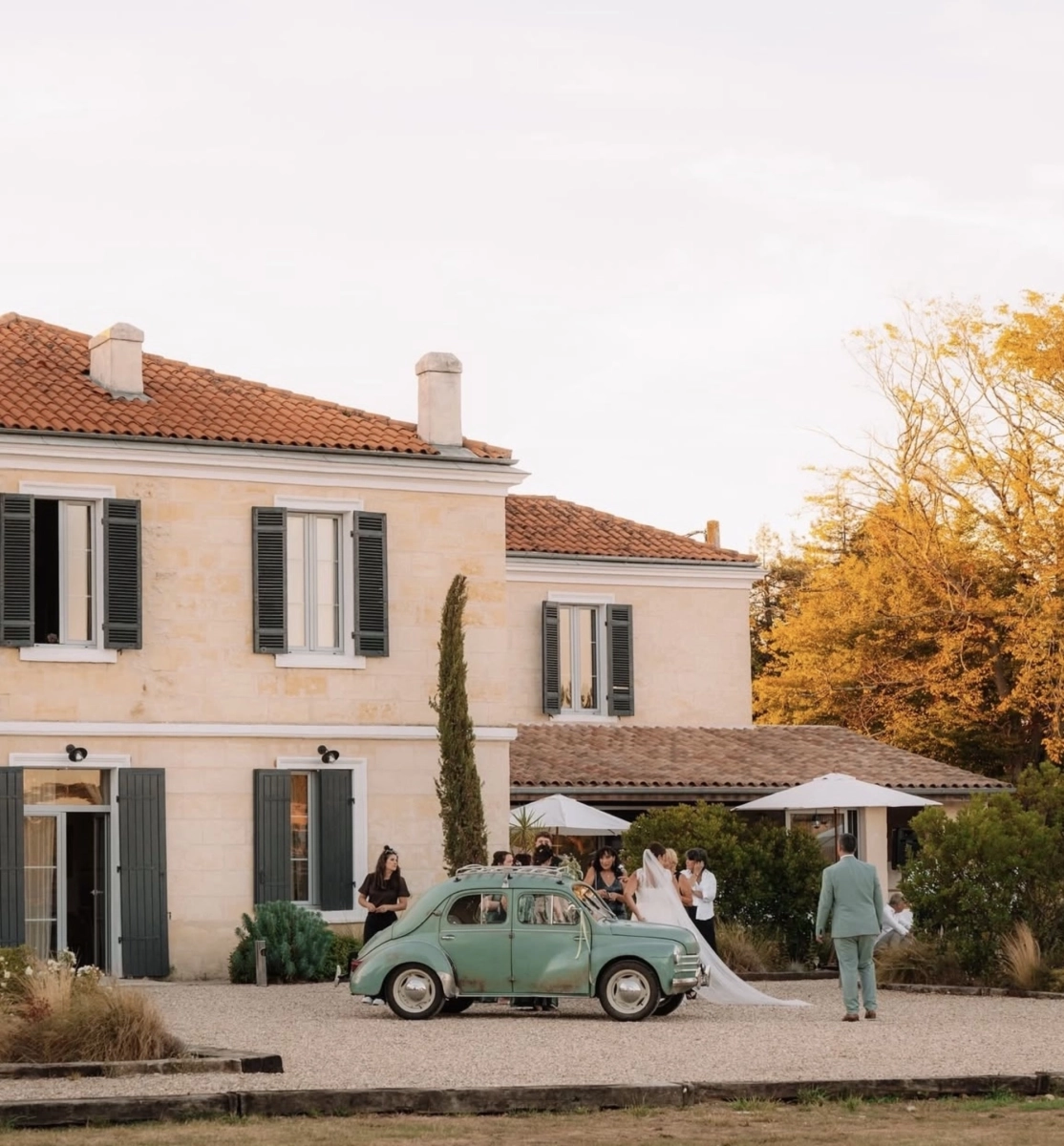 A wide outdoor shot taken in front of a two-storey French stone manor house with terracotta roof tiles and dark grey shutters, set on a gravel courtyard. A vintage mint-green Fiat 4 (Renault 4 style) car with white ribbon decoration is parked in the foreground, acting as a wedding vehicle. A bride in a white gown with a long cathedral-length veil stands beside the car, surrounded by approximately four to five people including a groom or guest in a sage green suit walking toward the group. Several guests dressed in black are also visible nearby. Two large white parasols are set up to the right side, suggesting a cocktail hour area. The overall styling is relaxed and classic with a French countryside aesthetic. Potential venue feature image.
