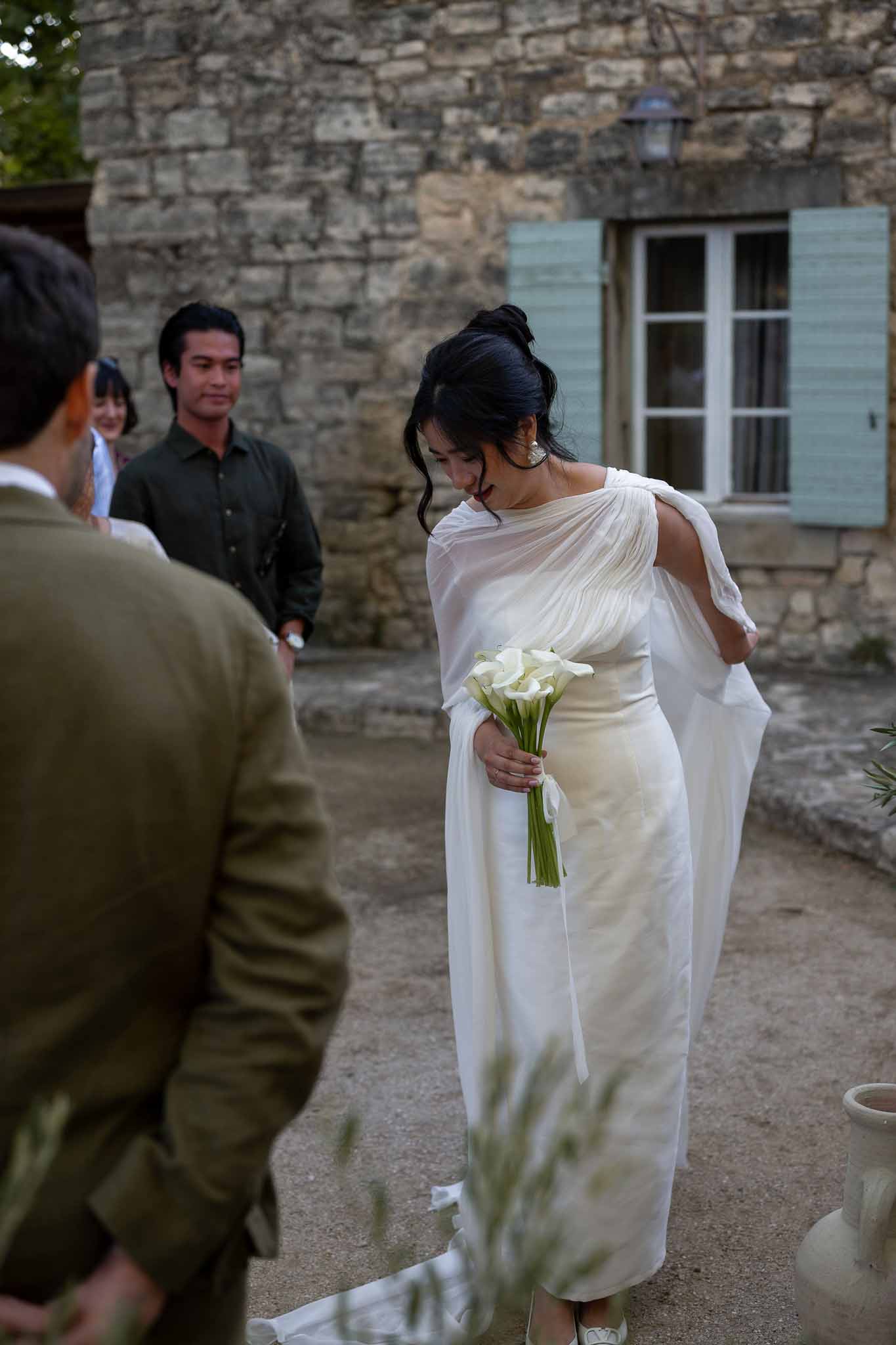 Bride in white gown standing at olive-branch decorated altar with calla lily bouquet