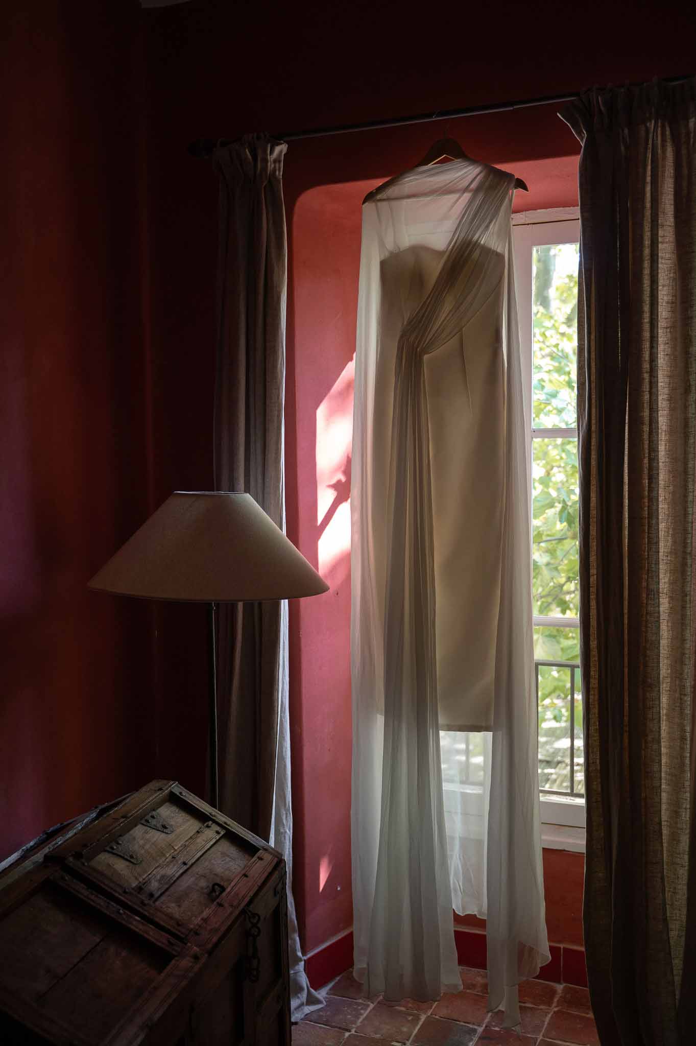 Bride in sheer short dress standing at bedroom window with natural light