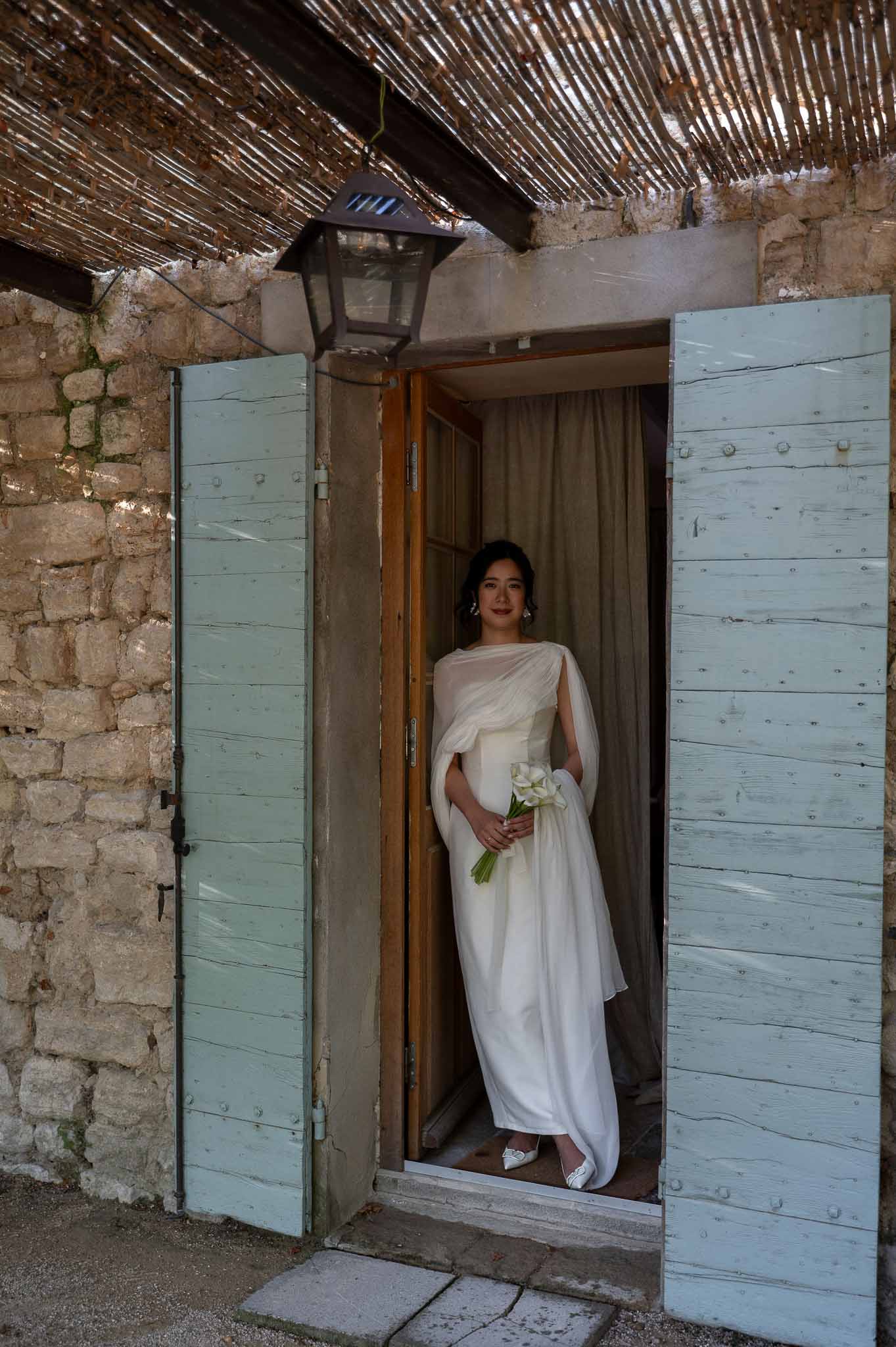 Bride in elegant Odd Muse gown standing at vintage wooden front door of Provençal bastide