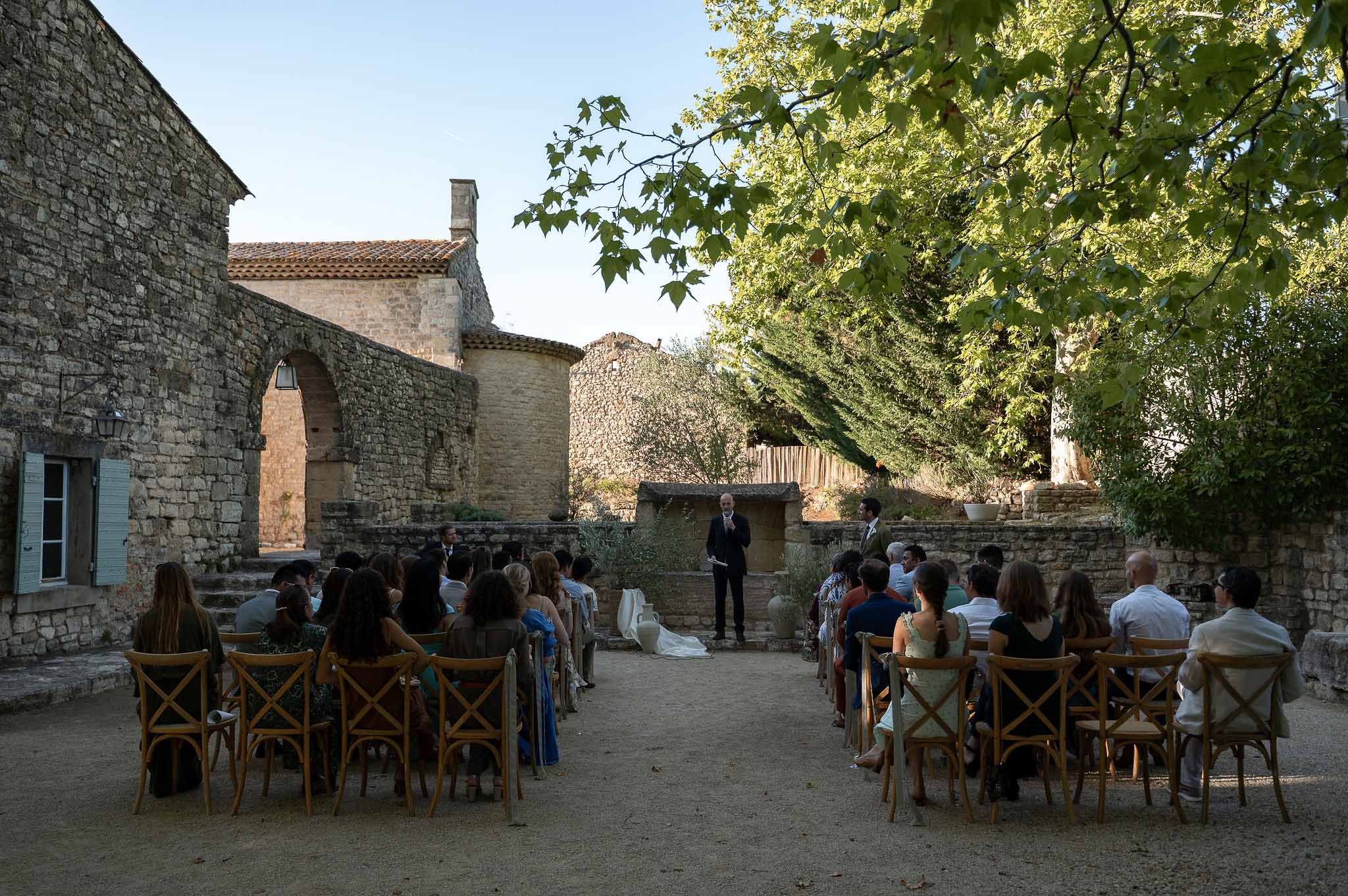 Wedding guests seated in wooden chairs at outdoor ceremony in garden at Notre-Dame de Conil