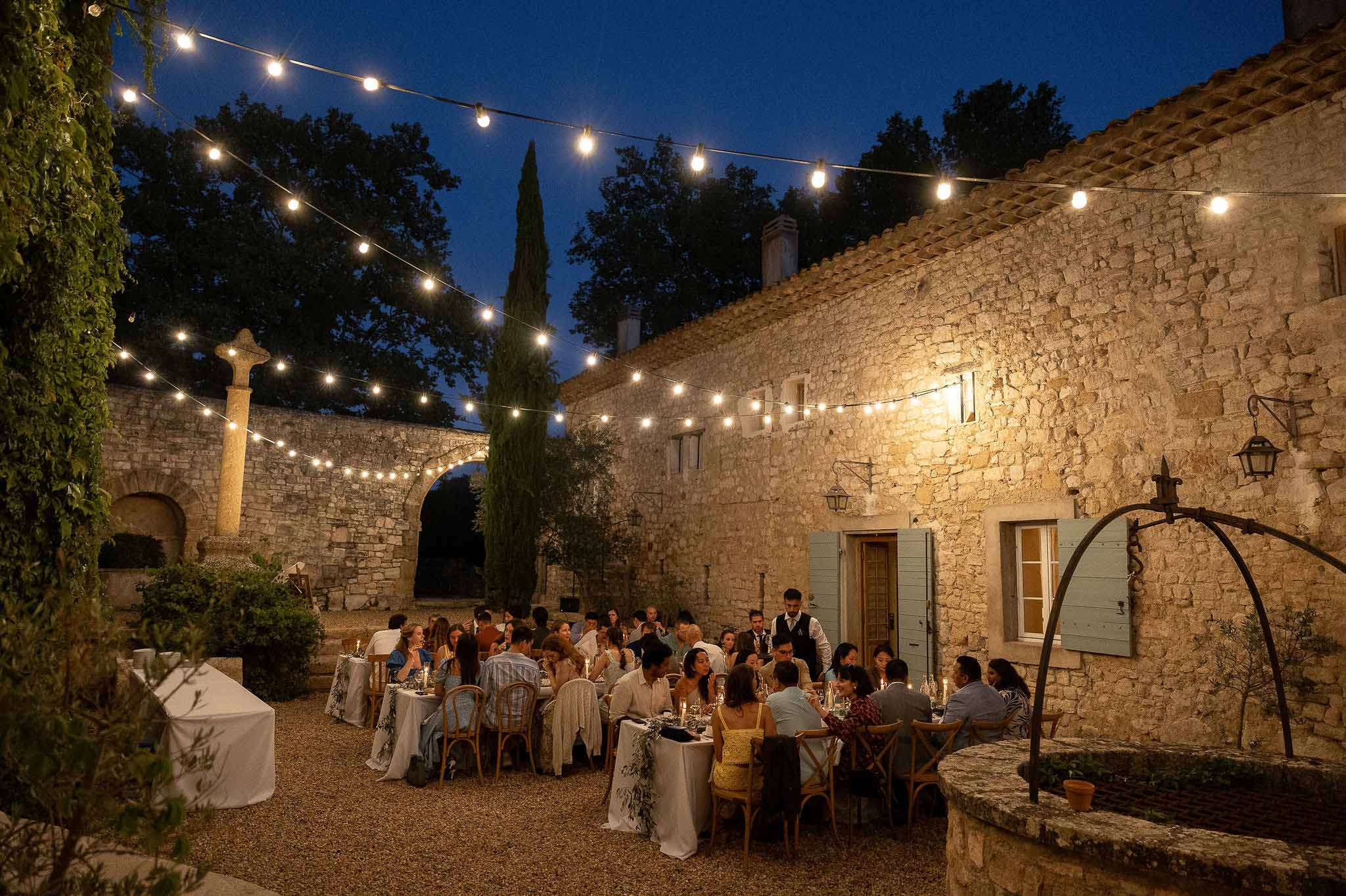 Wedding guests dining at long tables under warm string lights at outdoor garden reception