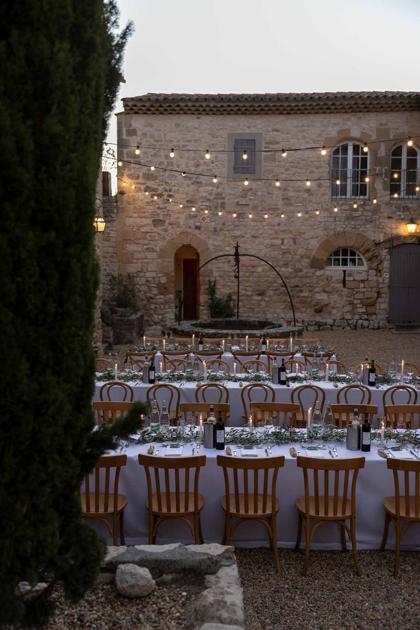 Long reception table under warm string lights with wooden chairs and olive branch styling