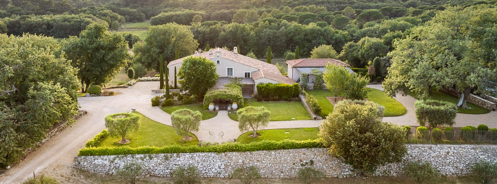 Aerial wide shot of a Provençal mas-style property surrounded by dense woodland, captured in warm daytime light. The main building features white-rendered walls and terracotta roof tiles, with a vine-covered entrance archway flanked by two large white ceramic urns. The grounds include a manicured lawn, clipped topiary hedges, cypress trees, and several olive trees arranged around a gravel courtyard and driveway. A dry-stone wall borders the front of the property, and a secondary building with a curved terracotta roof is visible to the right. No people are present in the image. Potential venue feature image.