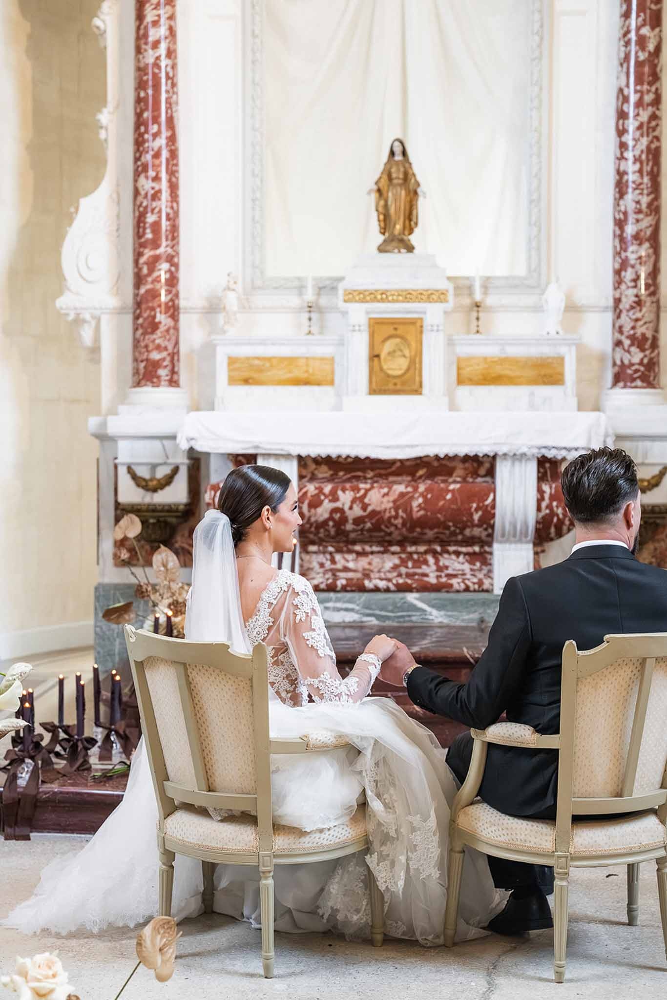 Couple seated holding hands at chapel altar at Chateau du Terral from behind