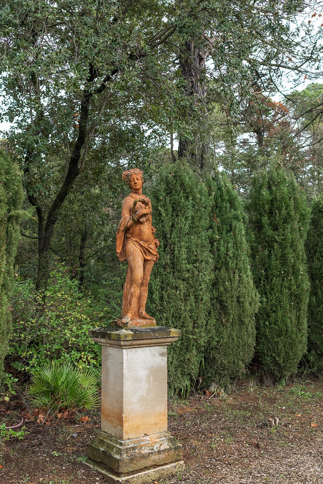 Weathered terracotta garden statue on plinth beside cypresses at Chateau du Terral