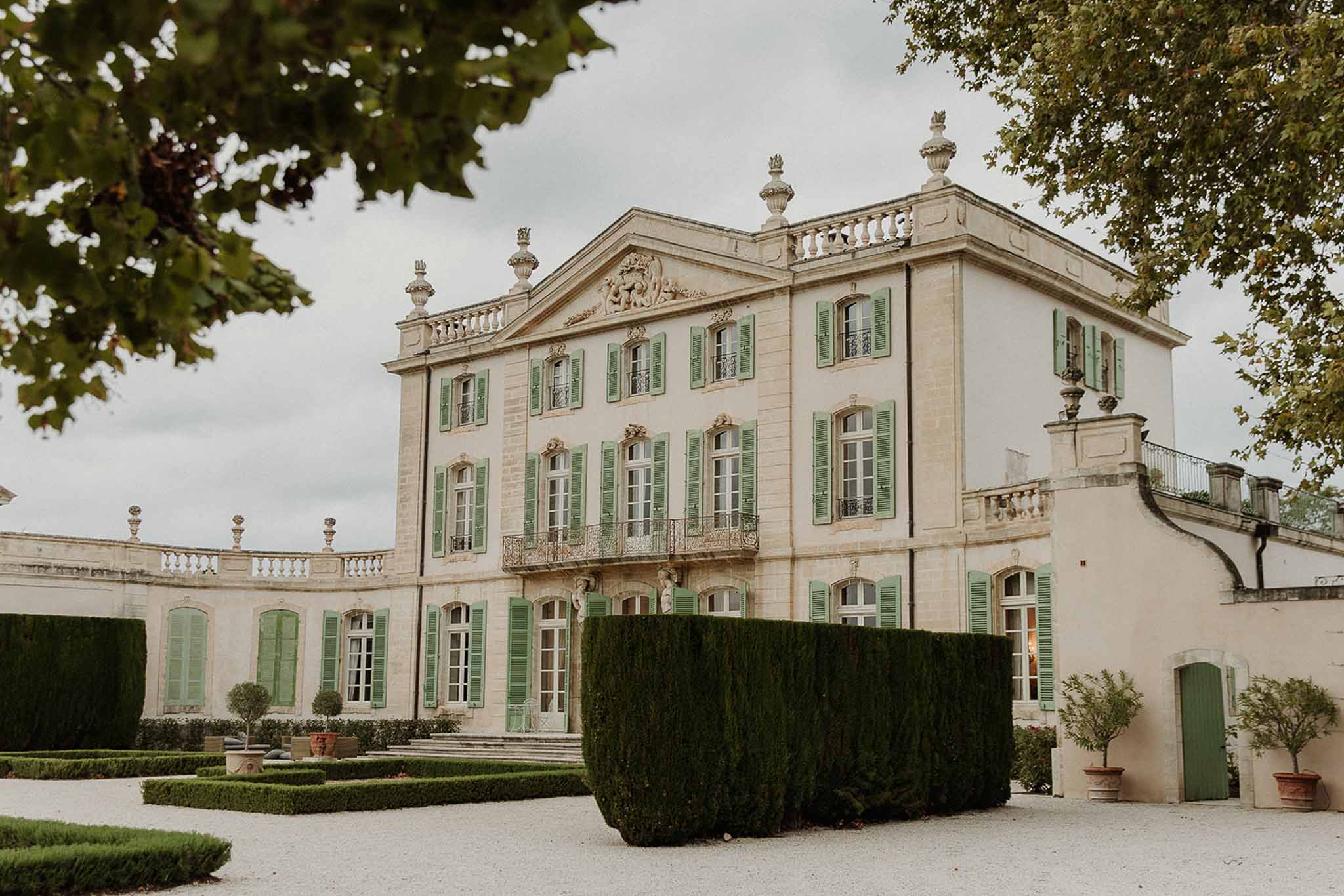 An exterior wide shot of a French classical château featuring cream limestone architecture with sage green shutters across multiple floors, ornate stone balustrades, decorative finials, and carved pediment detailing above the central facade. The forecourt features a gravel courtyard with formally clipped dark green topiary hedges, low box hedging parterre sections, and potted citrus trees flanking the entrance steps. No people are visible in the frame; the image functions purely as a venue exterior shot captured from a low angle with tree branches framing the upper corners. The overall architectural style is 18th-century French classical. Potential venue feature image.