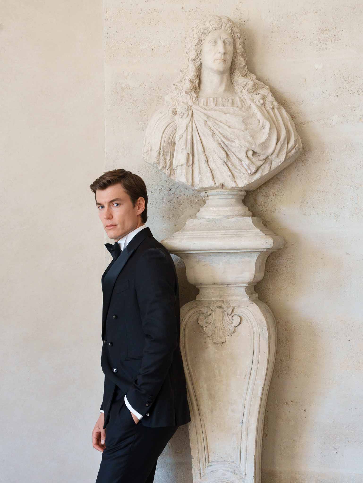 Groom in tuxedo posing beside a stone statue at the chateau