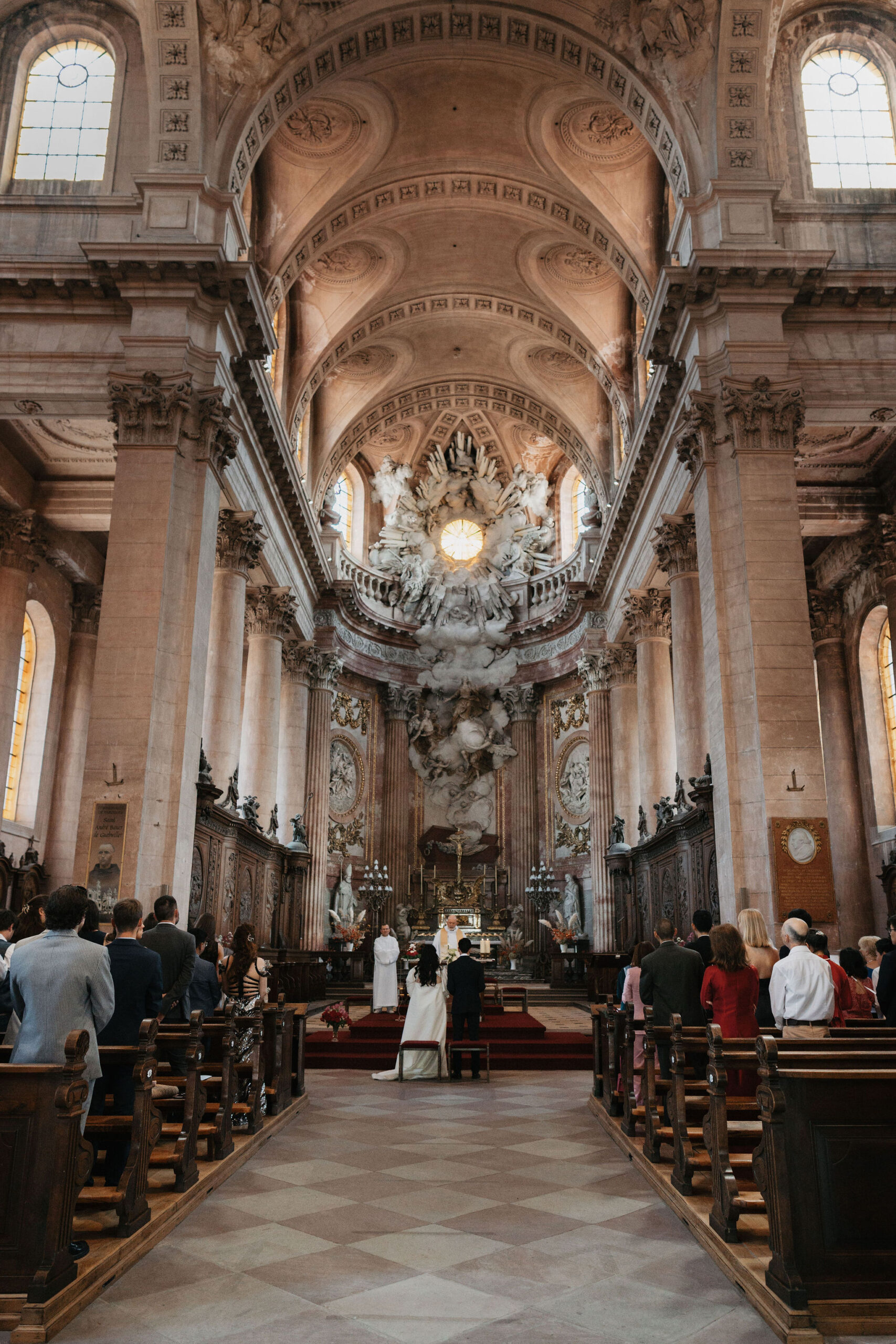 A religious wedding ceremony taking place inside a grand Baroque-style church or cathedral, shot from the back of the nave in a wide portrait orientation. The couple stands at the altar at the far end of the aisle — the bride in a long white gown and the groom in a dark suit — facing two officiants dressed in white liturgical robes. Approximately 40–60 guests are seated in dark wooden pews on either side of the central aisle, with a deep red carpet runner leading to the raised altar area. The altar is flanked by tall candelabras and small floral arrangements in warm orange and red tones, and the apse features an elaborate white sculptural group depicting figures in clouds, centered on a glowing circular element. The church interior is characterized by barrel-vaulted ceilings with ornate coffered detailing, tall arched windows, Corinthian pilasters, and carved wooden choir stalls lining the nave walls. Potential venue feature image.