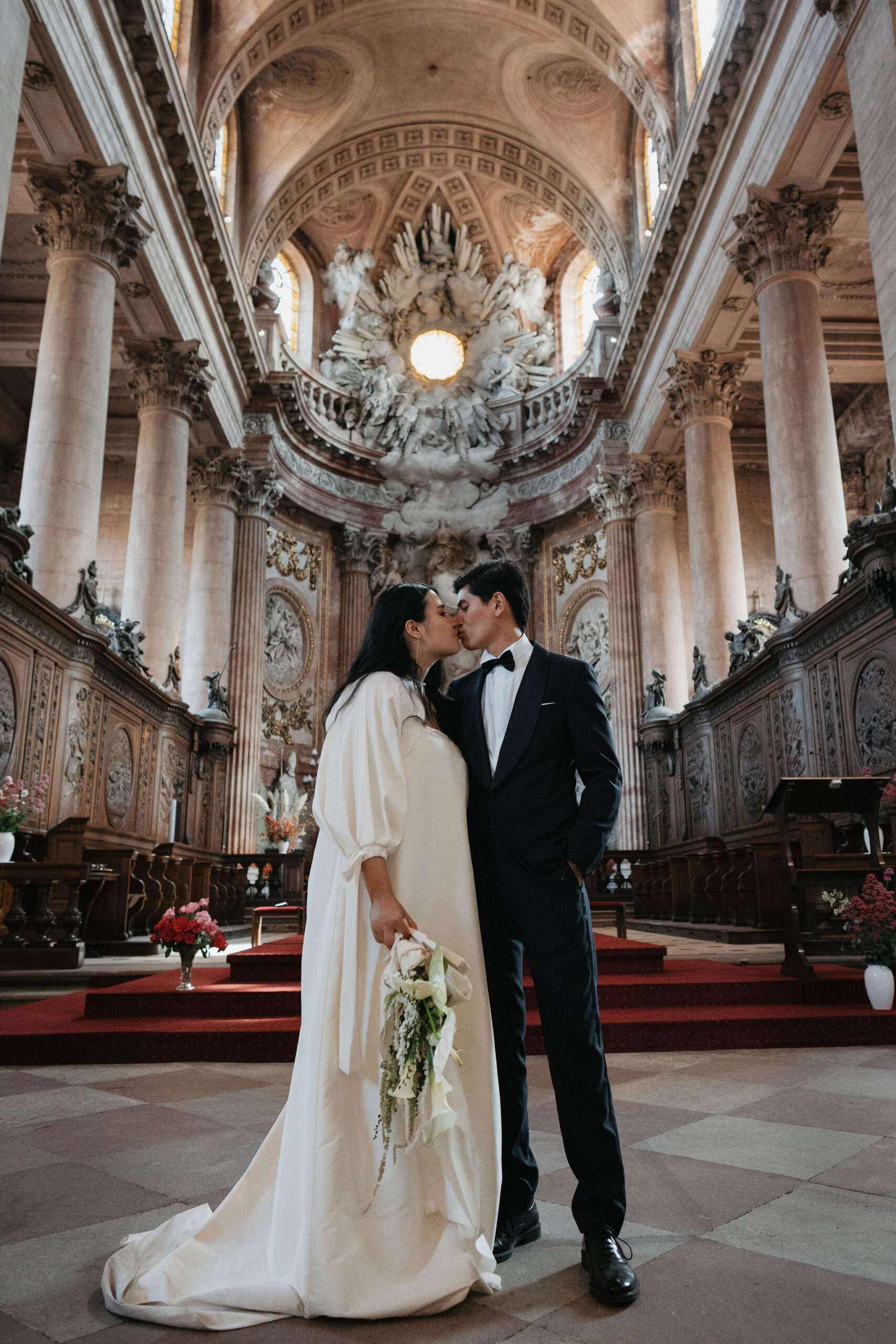 A couple shares a kiss during a portrait session inside a large baroque-style church or chapel, standing at the center of the nave with the ornate altar and carved wooden choir stalls visible behind them. The bride wears an ivory long-sleeve gown with voluminous bishop sleeves and a floor-length train, holding a trailing bouquet of white calla lilies, roses, and cascading greenery with a long white ribbon. The groom wears a black tuxedo with a white dress shirt and black bow tie. The interior features tall stone columns, elaborate sculptural ornamentation on the altar including white figures set against a gilded backdrop, red carpet on the chancel steps, and arrangements of red and pink flowers placed near the pews. The shot is a full-length wide portrait framed to include the full height of the vaulted ceiling, emphasizing the scale of the architectural space.