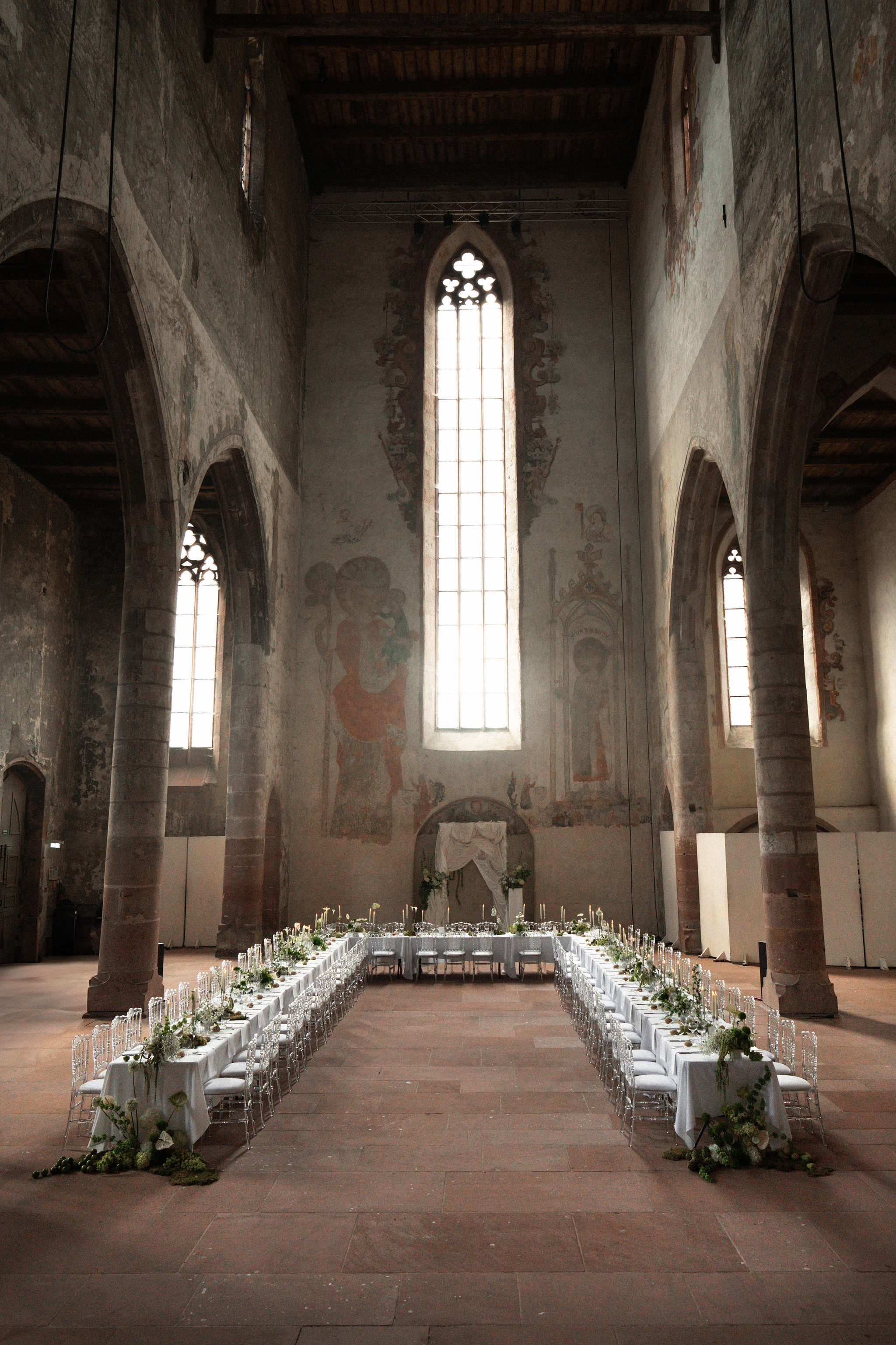A wide-shot image of a wedding reception setup inside a converted medieval church or abbey with soaring stone walls, tall Gothic-arched windows, exposed wooden ceiling beams, and large-scale faded frescoes depicting religious figures. Two long banquet tables draped in white linen run the full length of the nave, flanked by clear acrylic Chiavari chairs with white seat pads. The tables are decorated with trailing greenery runners featuring lush foliage and white blooms, slim taper candles in gold holders, and additional floral clusters placed on the stone floor at the table ends. A head table is set perpendicular at the far end in front of a draped fabric arch or altar feature with floral arrangements. The overall decor palette is white and green with a classic, minimalist approach against the raw historic architecture. No people are present — the room is fully set and empty. Potential venue feature image.