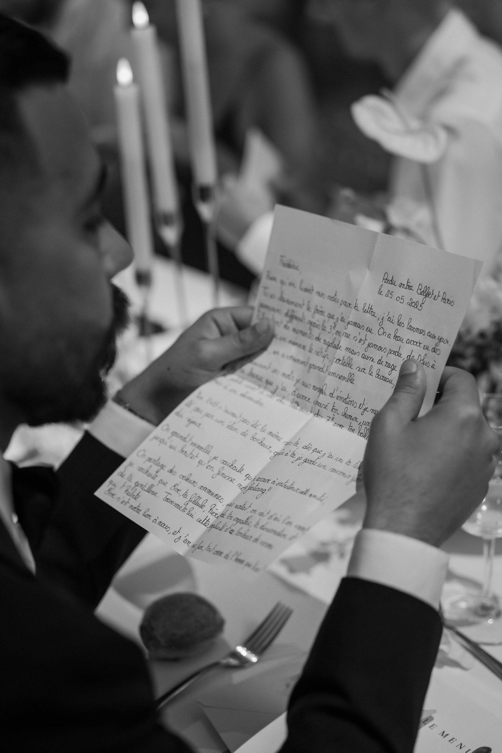 A black-and-white close-up portrait of a man in a dark suit and dress shirt reading a handwritten letter during what appears to be a wedding reception dinner. The letter is written in French and dated 25.05.2025, with the heading referencing 'Belfort et Paris.' The table setting is visible in the background and foreground, including a fork, white plate, a wine glass, and a printed menu card. Tall taper candles are lit in the background, suggesting an indoor evening reception. The shot is taken from over the man's shoulder, focusing on the handwritten pages he holds.