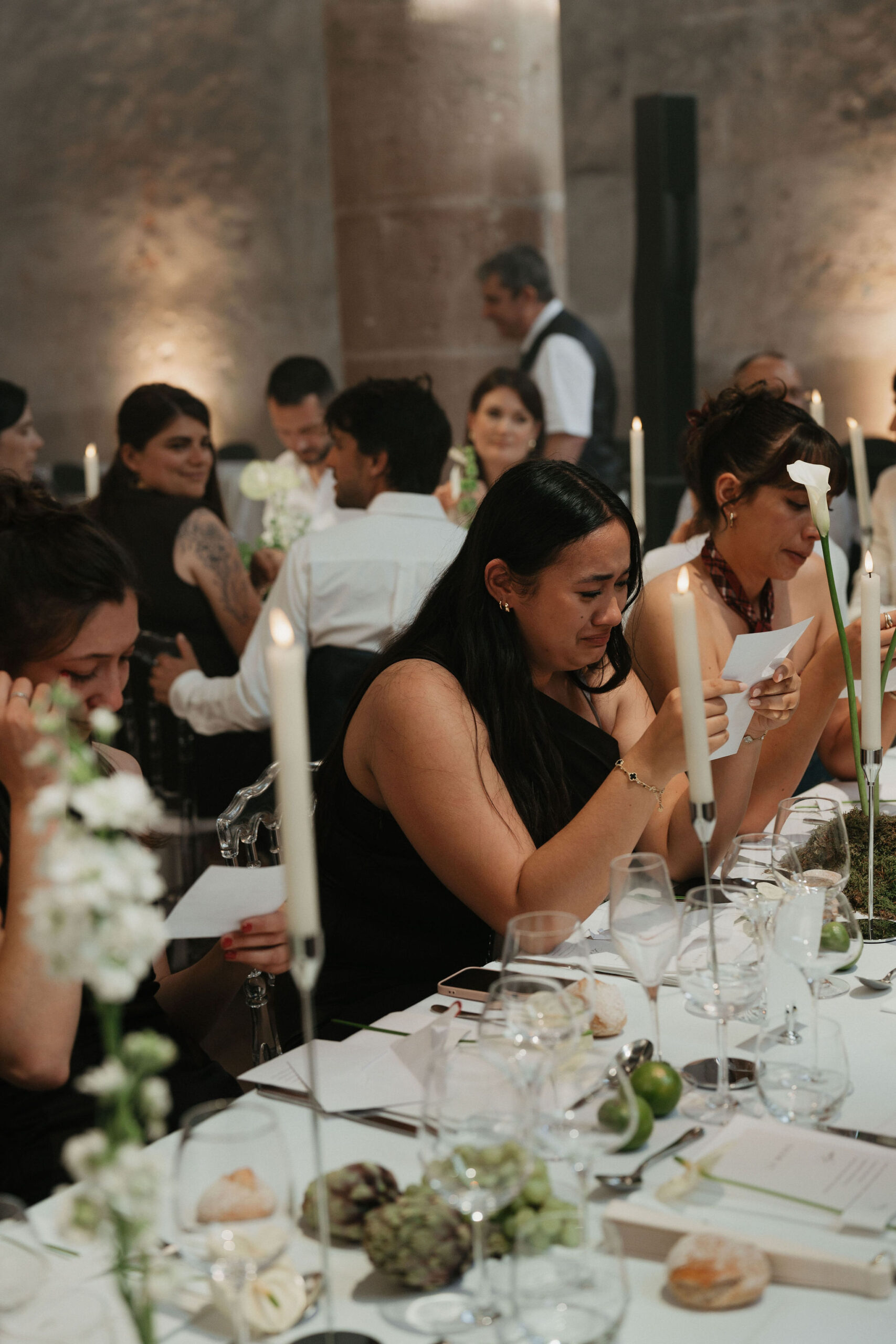 Indoor wedding reception dinner scene with approximately 10–12 guests seated and standing at a long dining table with a white linen tablecloth. A woman in a black one-shoulder dress in the foreground appears visibly emotional, wiping tears, while nearby guests also look moved, some holding small cards — suggesting a speech or vow reading is taking place. The table is decorated with tall white taper candles in slim holders, white ranunculus and small white blooms in low arrangements, single white calla lily stems, green apples, artichokes, and what appears to be a moss runner, creating a natural, modern-organic decor palette. Crystal glassware, white place cards, and a laid menu are visible on the table. The venue features large stone or marble columns and warm uplighting, indicating a historic indoor hall or château reception room. A staff member in a waistcoat is visible in the background. Medium-distance candid shot with shallow depth of field.