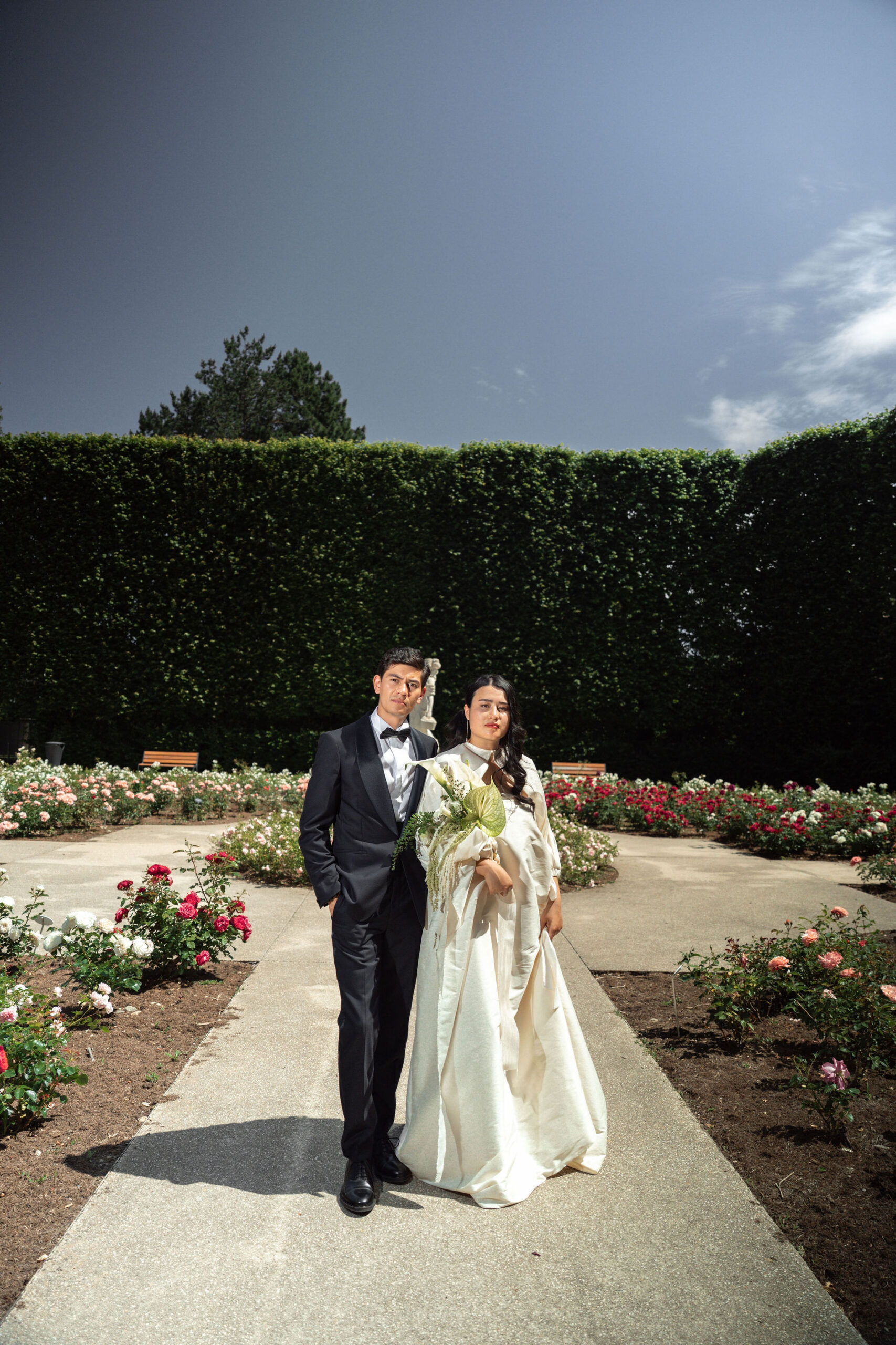A couple portrait taken outdoors in a formal rose garden, with the bride and groom standing together on a paved central pathway and facing the camera. The groom wears a black tuxedo with a white dress shirt and black bow tie, while the bride wears a structured ivory long-sleeve gown with a high collar and full skirt; she carries a loose bouquet featuring white anthurium blooms and large tropical green leaves. The garden beds on either side of the path are planted with red, pink, and white roses in full bloom, and a white classical statue is visible in the background flanked by wooden benches. The backdrop features tall, neatly clipped hedgerows forming a formal enclosure. The shot is a full-length couple portrait taken from a slightly low angle with a wide-angle lens.