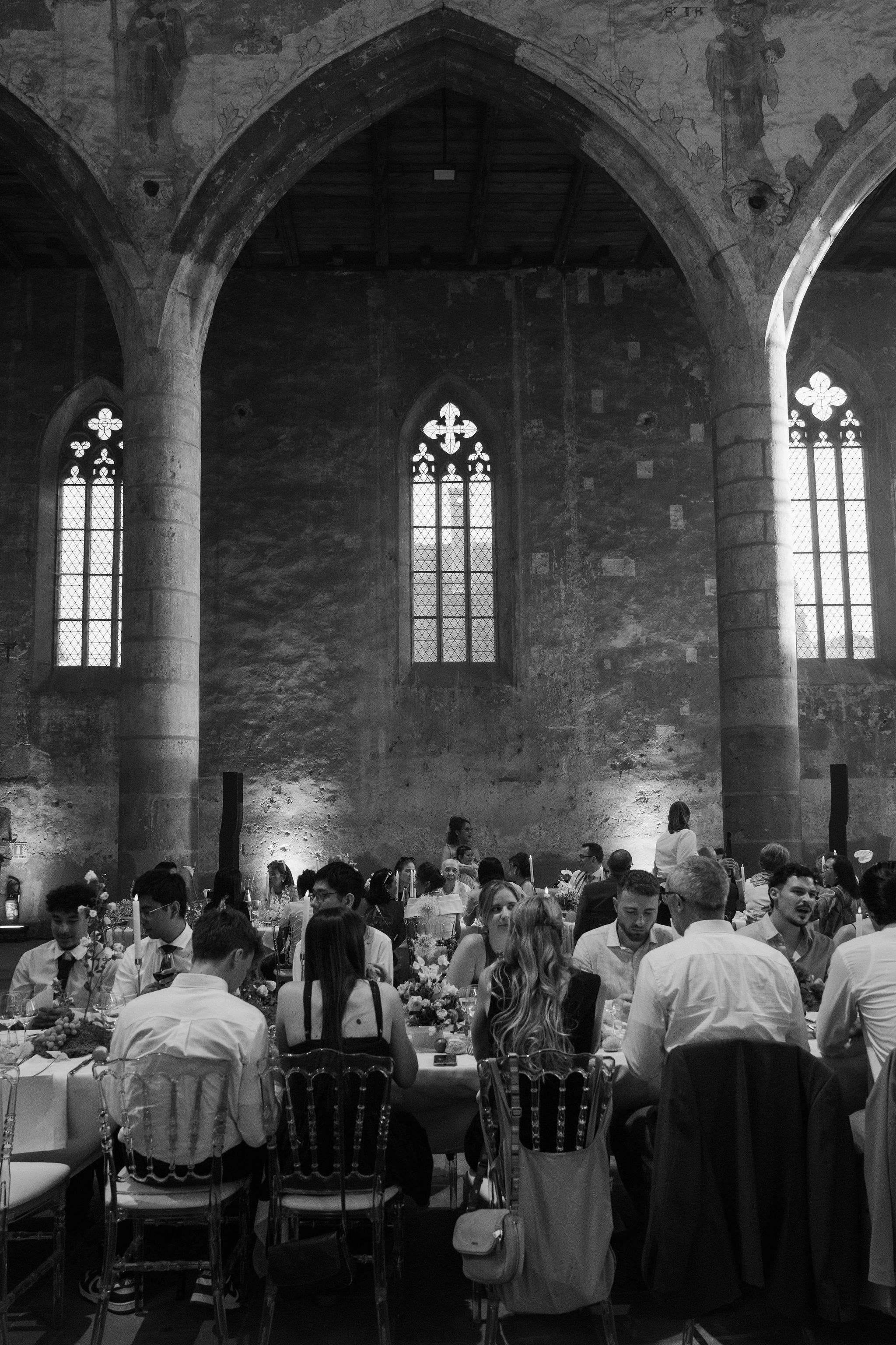 This black-and-white image shows a wedding reception dinner taking place inside a large medieval Gothic building, likely a converted abbey or chapel, with soaring pointed stone arches, tall lancet windows with quatrefoil tracery, exposed stone walls with traces of frescoes, and a wooden beam ceiling. Warm uplighting illuminates the lower sections of the stone walls, creating strong contrast against the bright windows. Approximately 30–40 guests are seated at long banquet tables set with candles, floral centerpieces of mixed blooms, and glassware, with additional guests visible deeper in the space. Guests are dressed in smart casual to formal attire, and Chiavari-style chairs are visible throughout. The wide-angle shot is taken from ground level near the back of the room, capturing both the architectural height and the full length of the reception setup. Potential venue feature image.