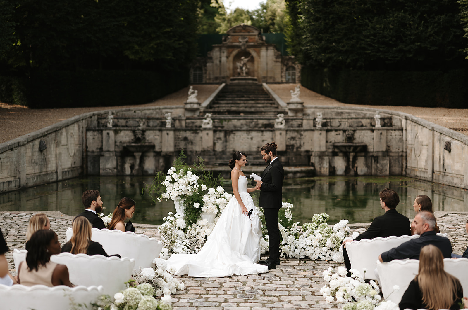 An outdoor wedding ceremony taking place on a cobblestone terrace in front of a large formal reflecting pool, with a grand stone staircase, classical statuary, and ornamental fountain structure serving as the backdrop. The bride wears a white strapless ballgown with a full skirt and cathedral-length train, while the groom is dressed in a black tuxedo with bow tie and glasses, holding paper vows as the couple faces each other. The ceremony is framed by large white urn arrangements overflowing with white peonies, hydrangeas, and lush greenery, with a ground-level floral carpet of white blooms — including baby's breath and hydrangeas — lining the aisle on both sides. Guests, dressed predominantly in black formal attire, are seated in rows of ornate white carved chairs on either side of the aisle; approximately 20–30 guests are visible. The overall design palette is strictly white and green, with a classic, architectural styling theme. This is a wide-shot taken from behind and slightly elevated above the guest seating, capturing the full ceremony setup, the couple at the altar, and the monumental formal garden architecture behind them. Potential venue feature image.